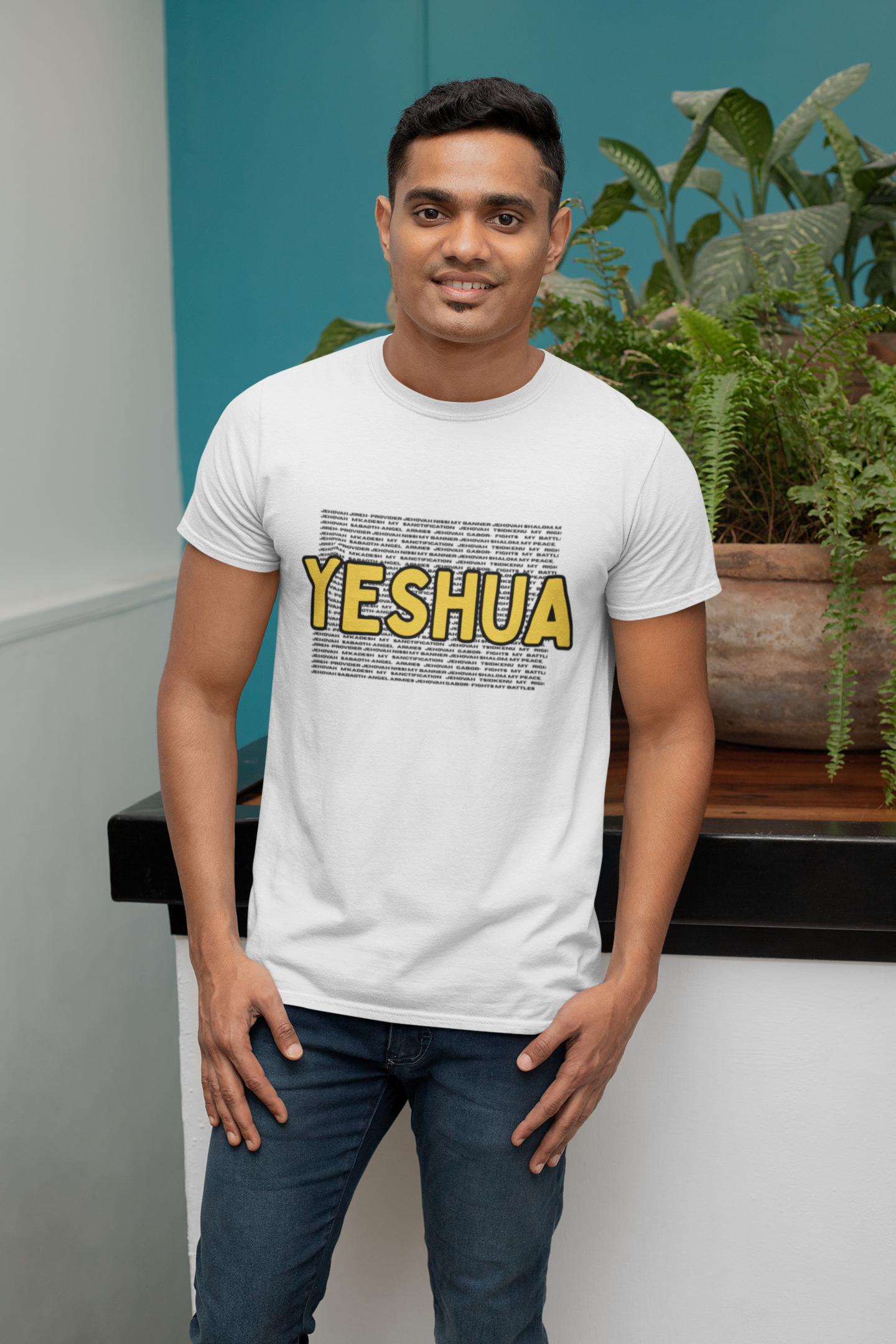 Christian T Shirt. Man wearing a white t-shirt with 'YESHUA' printed in yellow, standing indoors with plants in the background.